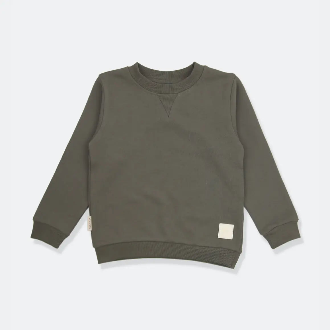 An olive green long-sleeve crewneck sweatshirt for a toddler, laid flat on a light gray background. It features a small cream-colored square patch on the lower right hem and a small fabric tag on the lower left sleeve.