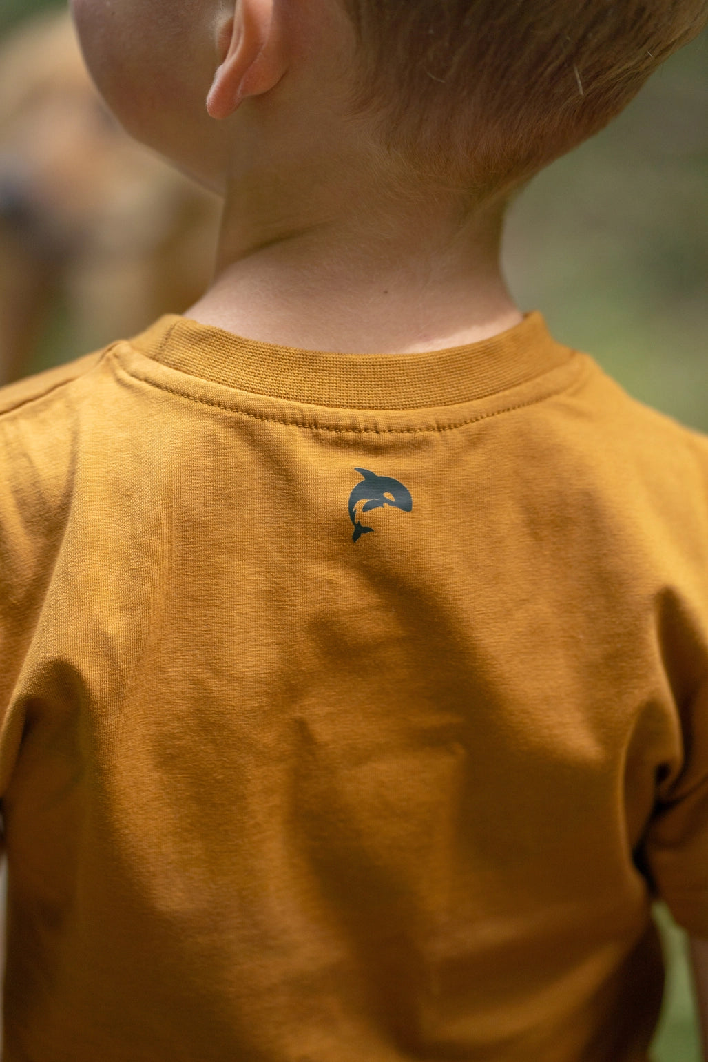 Close-up back view of a child wearing a mustard yellow t-shirt with a small dark blue dolphin logo on the upper back.
