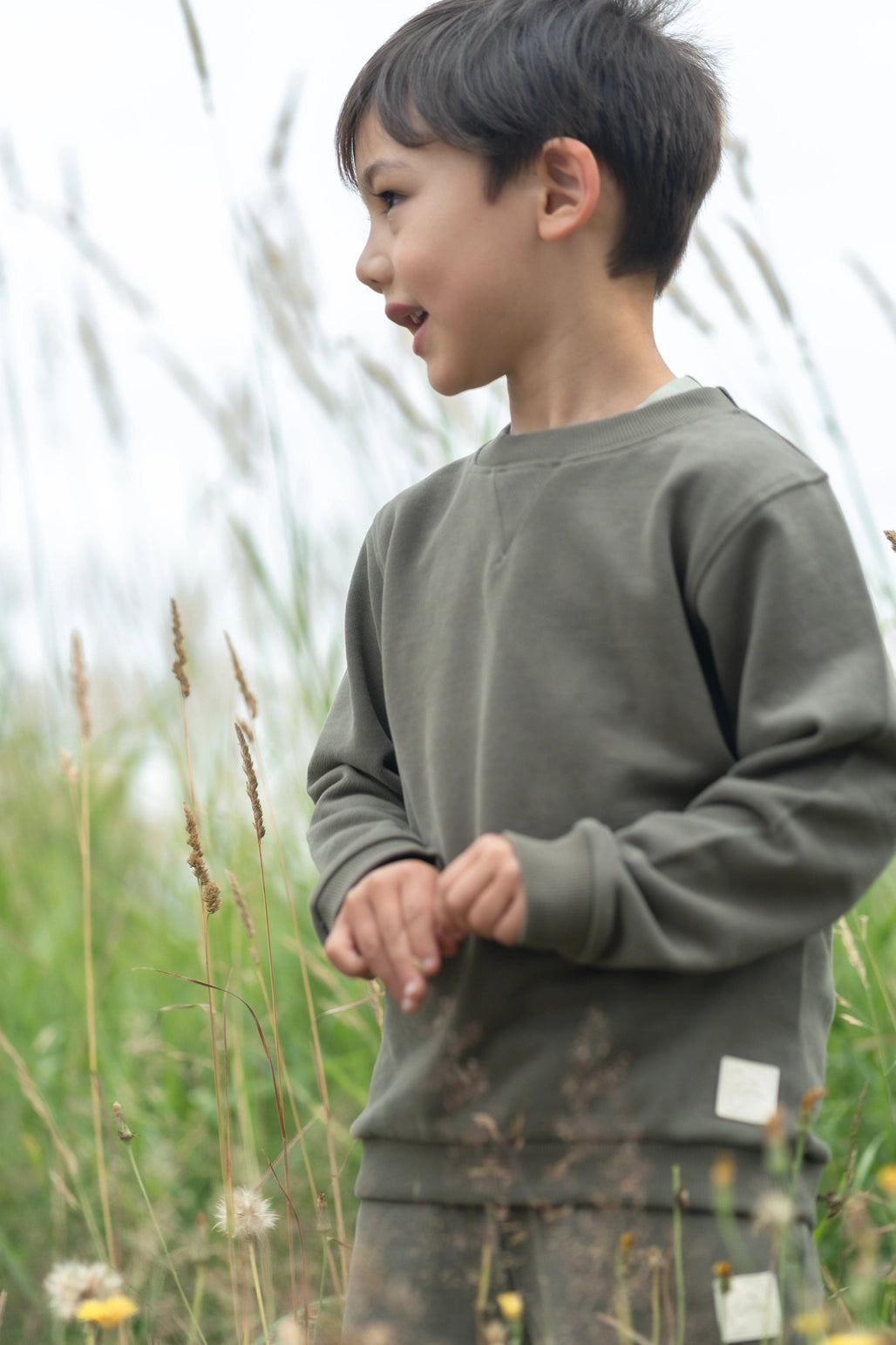 A young boy wearing an olive green crewneck sweatshirt and matching pants, standing in a grassy field.