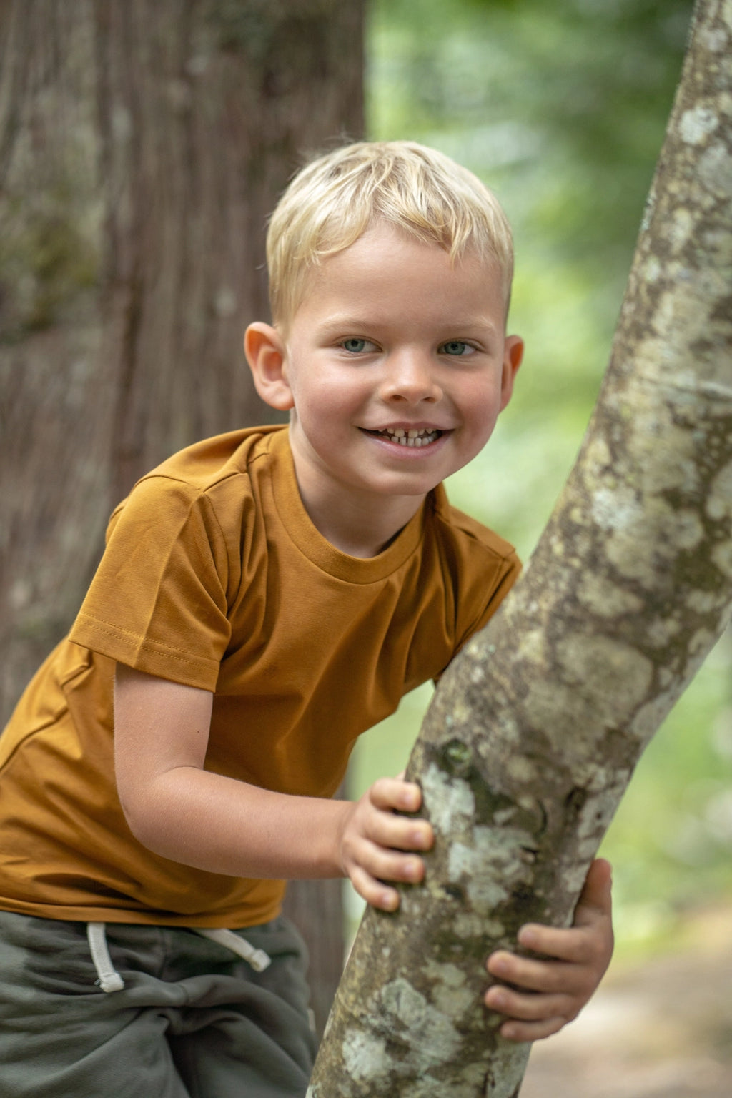 A young blonde boy smiling while holding onto a tree trunk outdoors in a forest setting, wearing a mustard yellow t-shirt and dark gray pants.