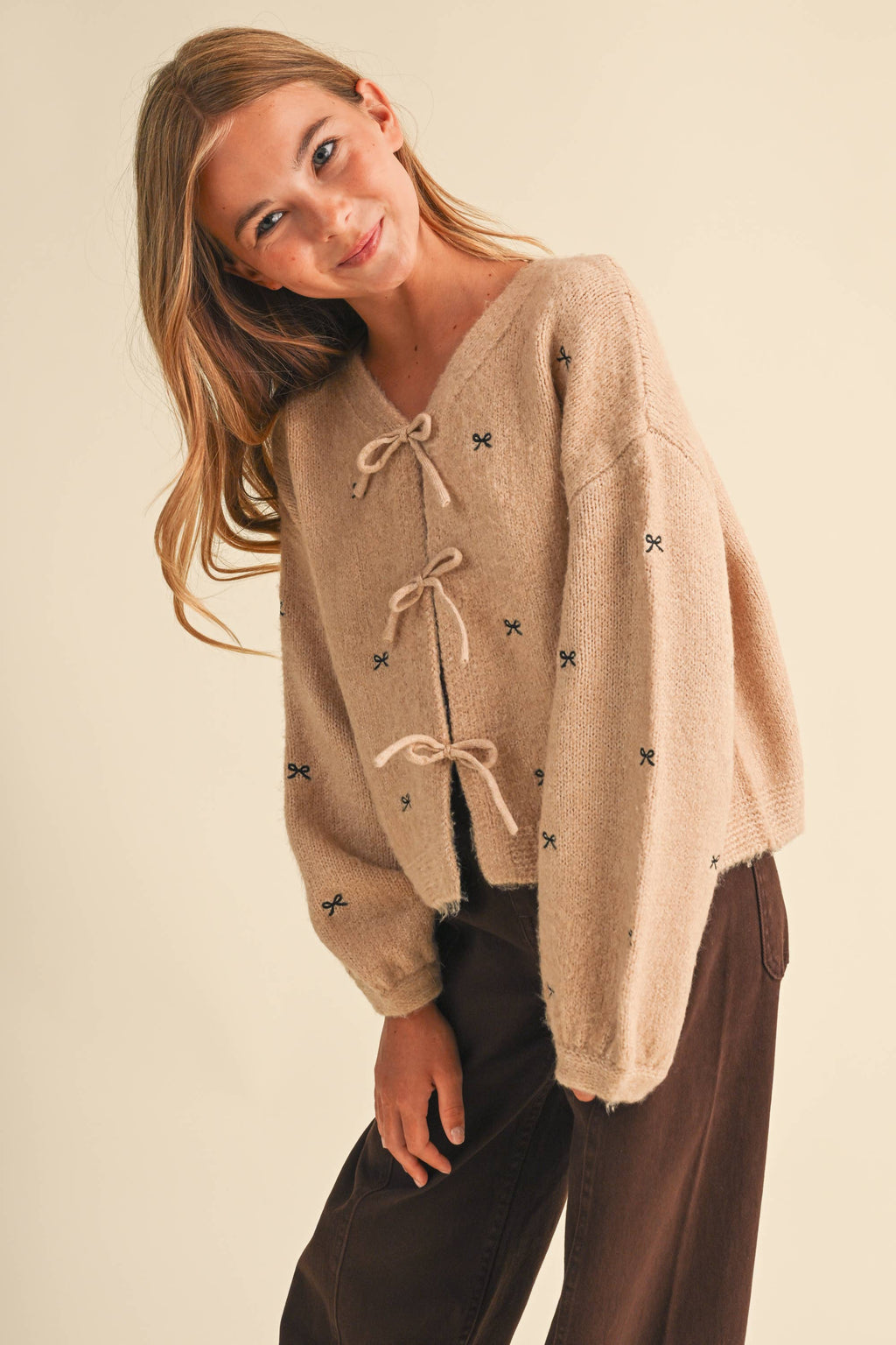 A young girl wearing a beige fuzzy knit cardigan with tie closures and small embroidered star details, paired with dark brown pants, posing against a solid light beige background.
