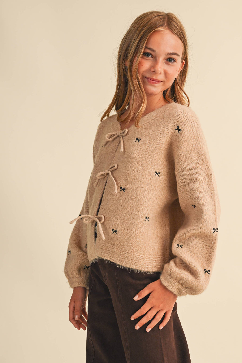 A young girl modeling a fuzzy beige knit cardigan with tie closures and small black embroidered star details, paired with dark brown pants, against a solid light beige background.