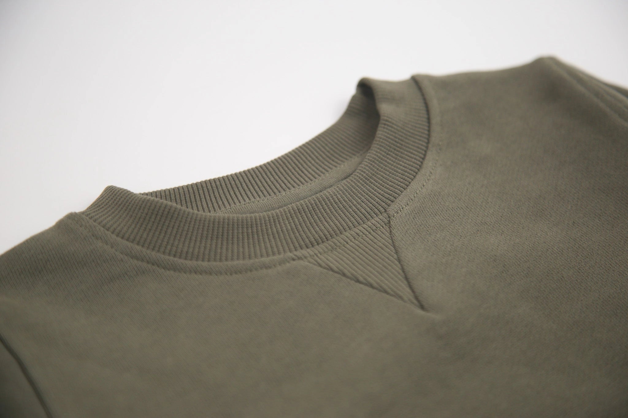 Close-up of a crewneck sweatshirt in olive green, showing the ribbed collar and shoulder seam against a light gray background.